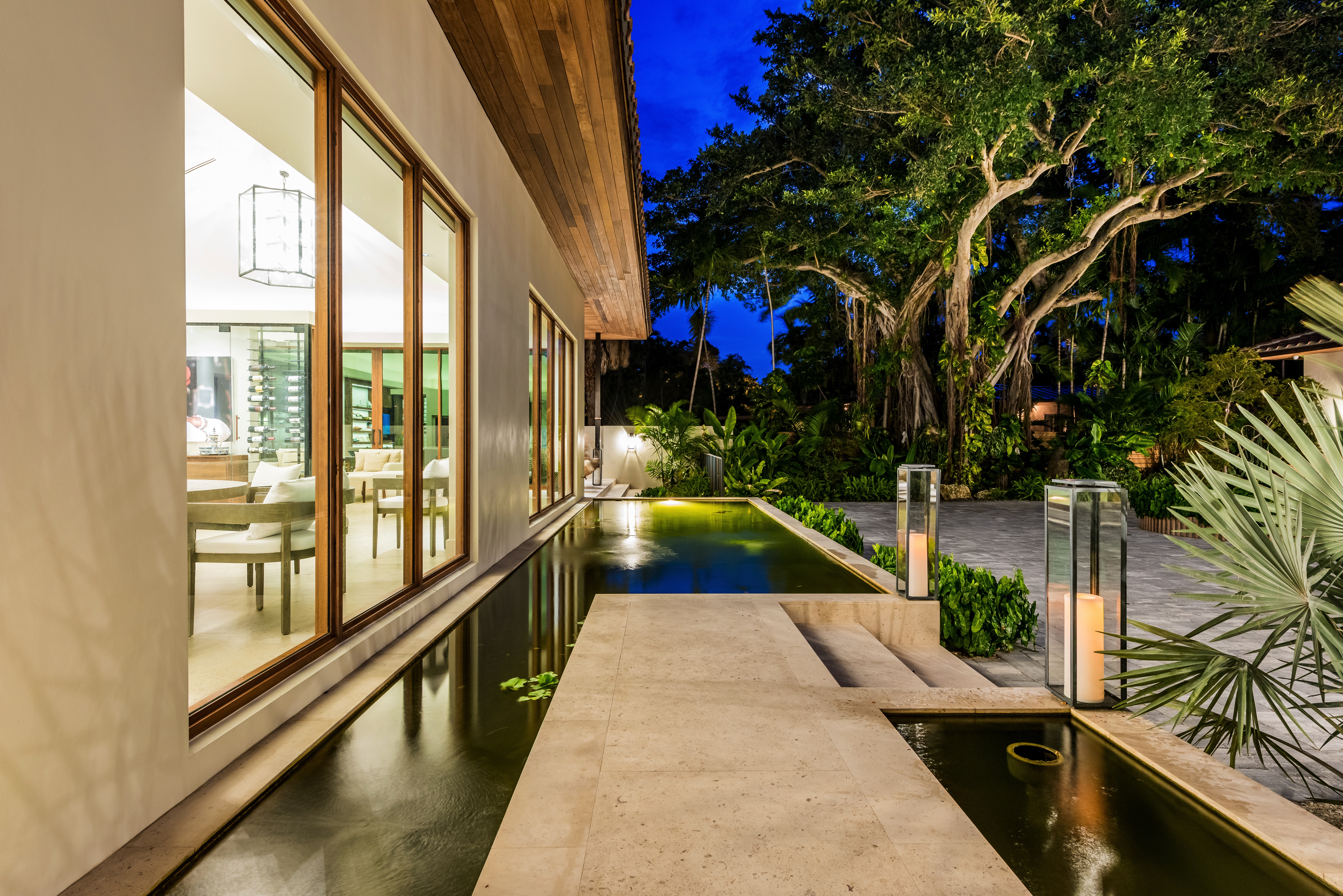 A stone walkway between lit reflecting pools beside a Miami villa at dusk, under a banyan tree
